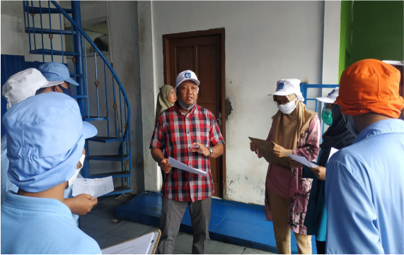 Seorang pria berbaju kotak-kotak berbicara kepada sekelompok orang yang mengenakan topi dan masker, di dalam ruangan dengan dinding berwarna biru dan hijau.