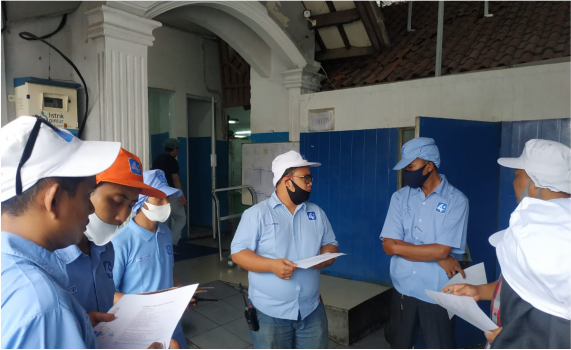 Sejumlah staf mengenakan seragam berwarna biru, berbincang dan memegang dokumen di area dalam gedung dengan dinding biru dan putih.