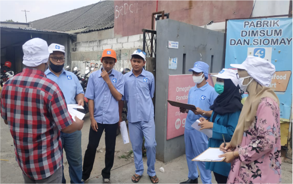 Sekelompok orang berbincang di luar pabrik, mengenakan seragam berwarna biru dan topi, serta masker, di depan pintu masuk pabrik dimsum.