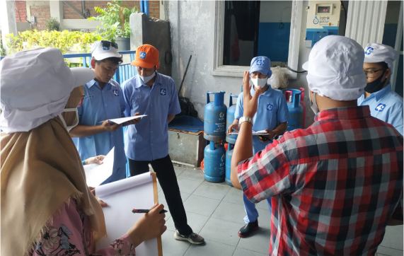 Meeting di lokasi kerja dengan beberapa peserta yang mengenakan seragam dan topi, sementara satu orang berdiri dan berbicara di depan kelompok.