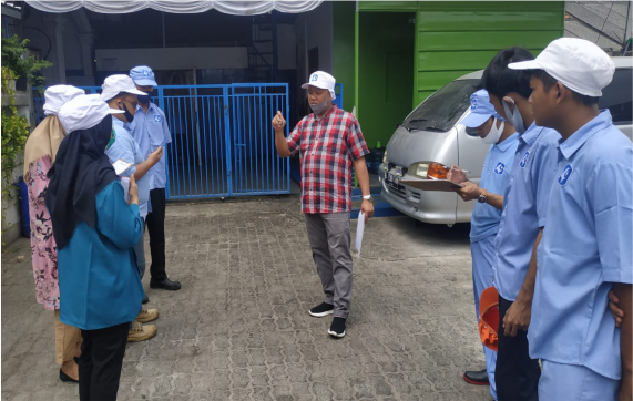 Seorang pria berbaju kotak-kotak berbicara di depan sekelompok orang yang mengenakan seragam biru dan topi putih di luar ruangan, sementara sebuah mobil terlihat di latar belakang.