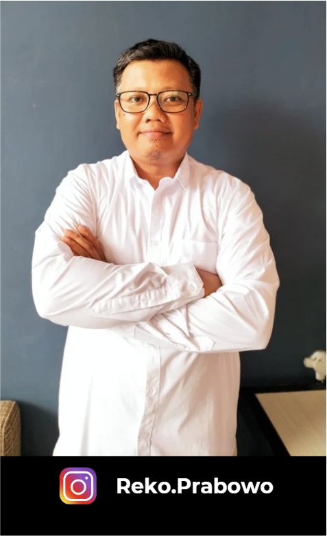 Seorang pria dengan kacamata dan baju putih, berdiri di depan latar belakang berwarna biru, dengan tangan bersedekap.